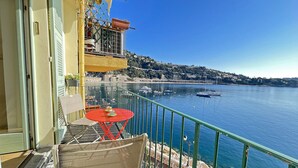Un balcó amb una taula i caires, amb vistes a un mar tranquil amb vaixells i un paisatge amb turons al fons.