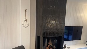 A modern living room with a dark wood fireplace, a comfortable armchair, a flat-screen TV, and a white shag rug.