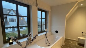 A modern bathroom with a bathtub, a toilet, and a shower area. There is a window with a view of a house outside.