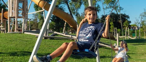 A child on a swing in a park with a playground structure in the background.