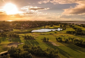 A golf course with multiple bunkers, a pond, and a sunset in the background.