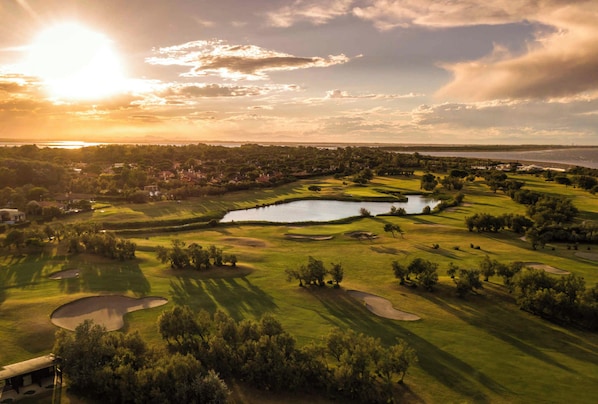 A golf course with multiple bunkers, a pond, and a sunset in the background.