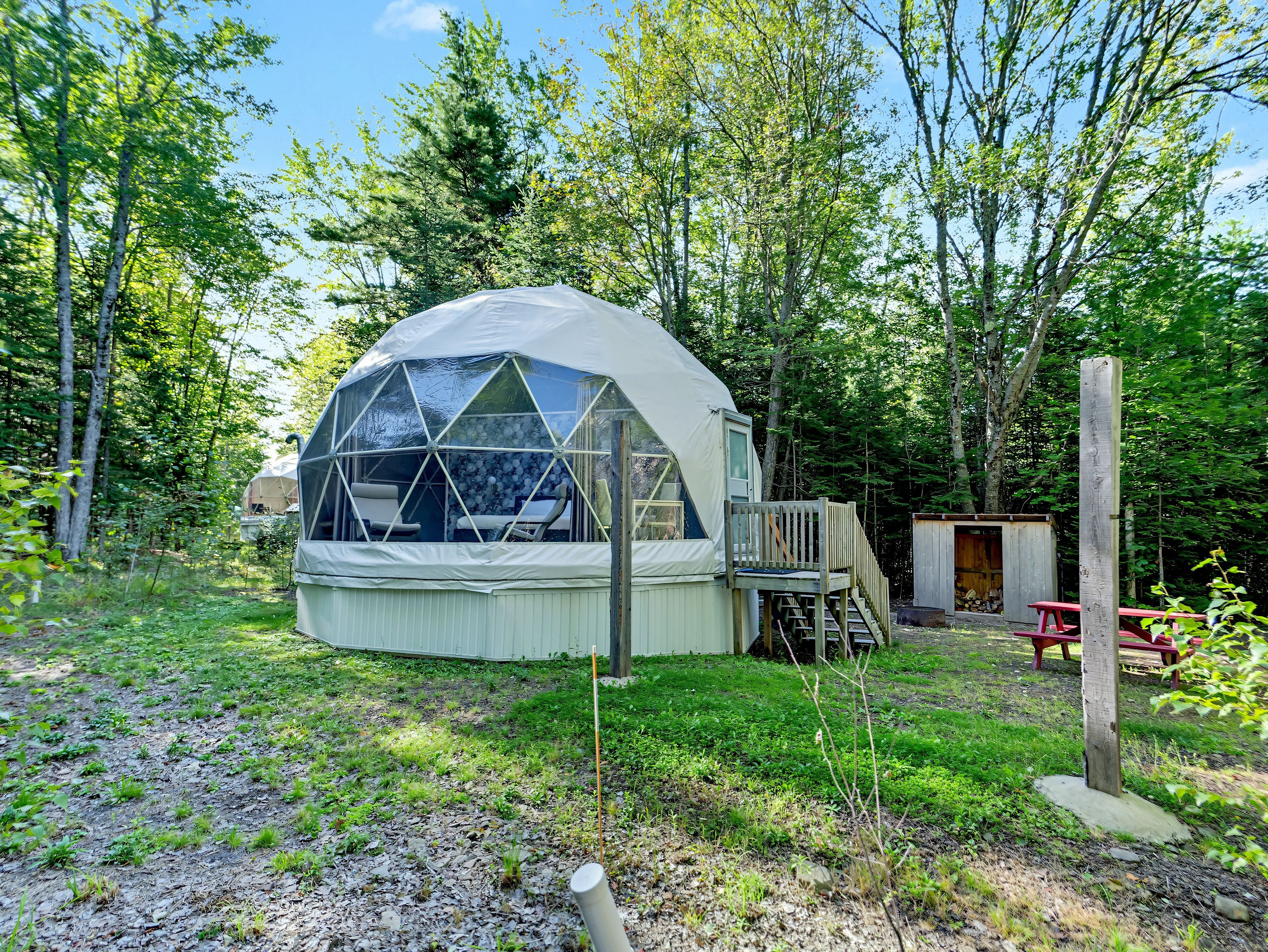Une structure en forme de dôme géodésique, dotée d’une terrasse et d’un petit abri, située dans une zone boisée.