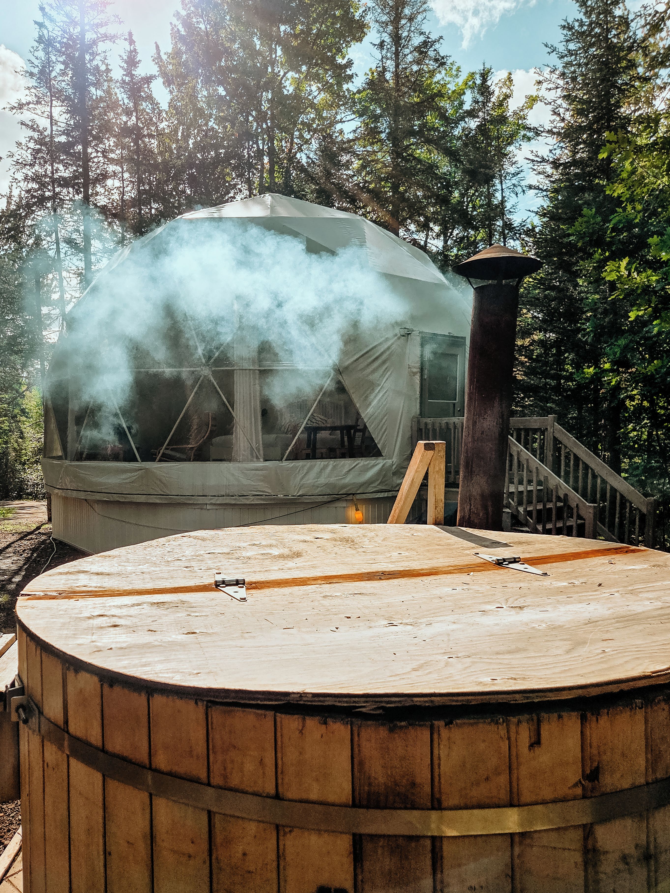 A hot tub with a wooden deck, surrounded by trees, and a smoke-emitting structure in the background.