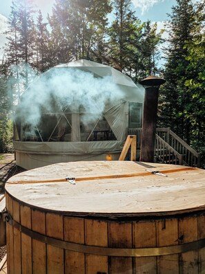 A hot tub with a wooden deck, surrounded by trees, and a smoke-emitting structure in the background. - Glamp Camp (Waterborough)