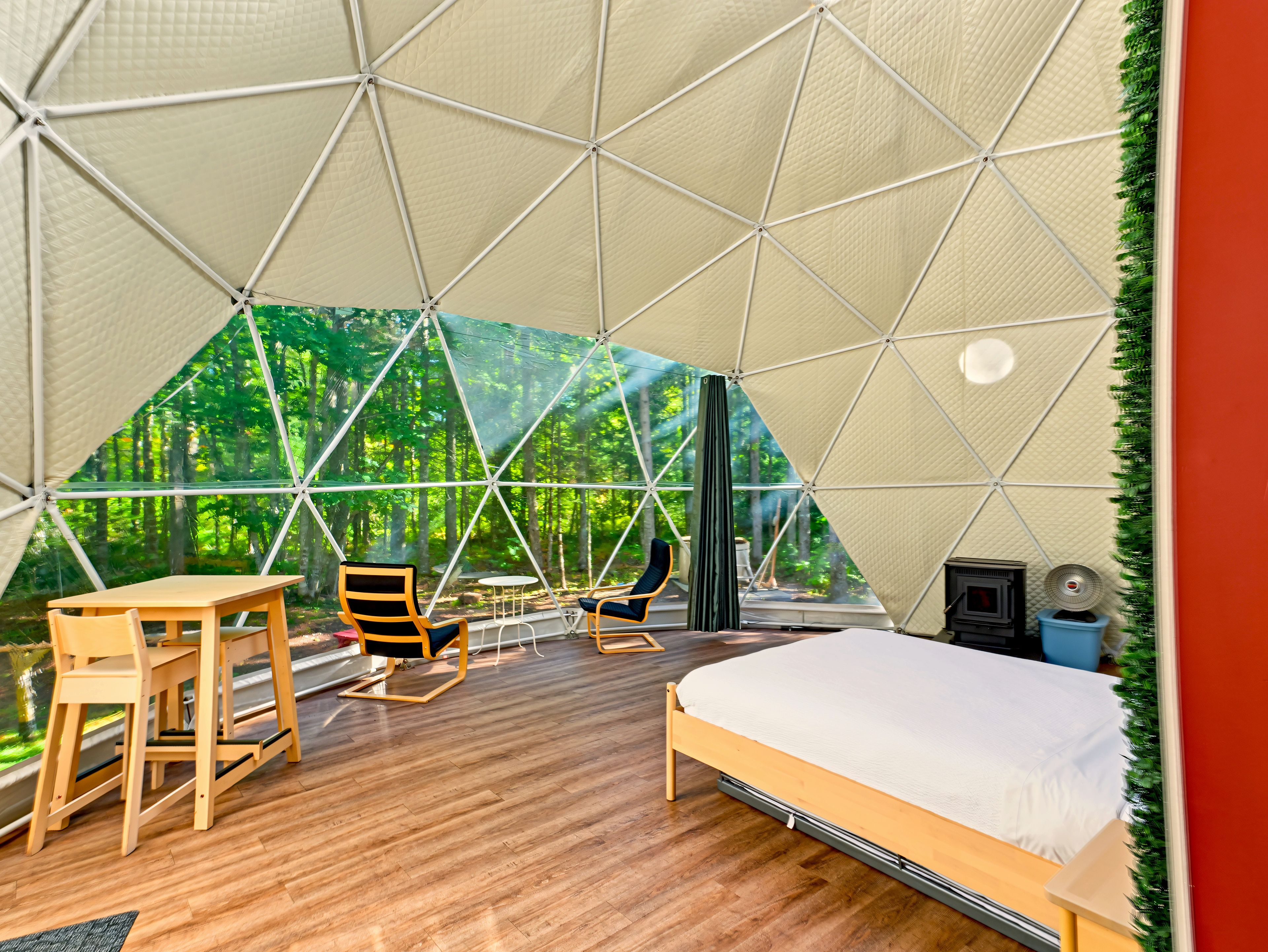 A geodesic dome room with a bed, two chairs, a table, and a view of a forest.