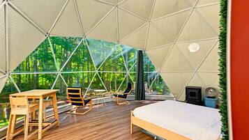 A geodesic dome room with a bed, two chairs, a table, and a view of a forest.