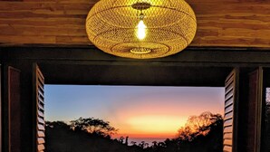 Uma área de refeições com mesa, cadeiras, luminária pendente e vista para o pôr do sol sobre a paisagem.