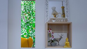 A room with a yellow chair, a small table, a window with a view of greenery, a shelf with decorative items, and a sign that says "I HOME". - 4u Apartment Athens! 2 Minutes From Metro Station (Nea Ionia)