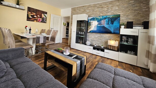 A modern living room with a flat-screen TV, a dining area with chairs and a table, and a kitchen in the background. - Bright and modern apartment on the ground floor in the center (Kassel)