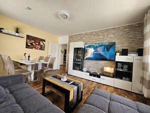 Ein modernes Wohnzimmer mit Flachbildfernseher, einem Essbereich mit Stühlen und Tisch sowie einer Küche im Hintergrund.