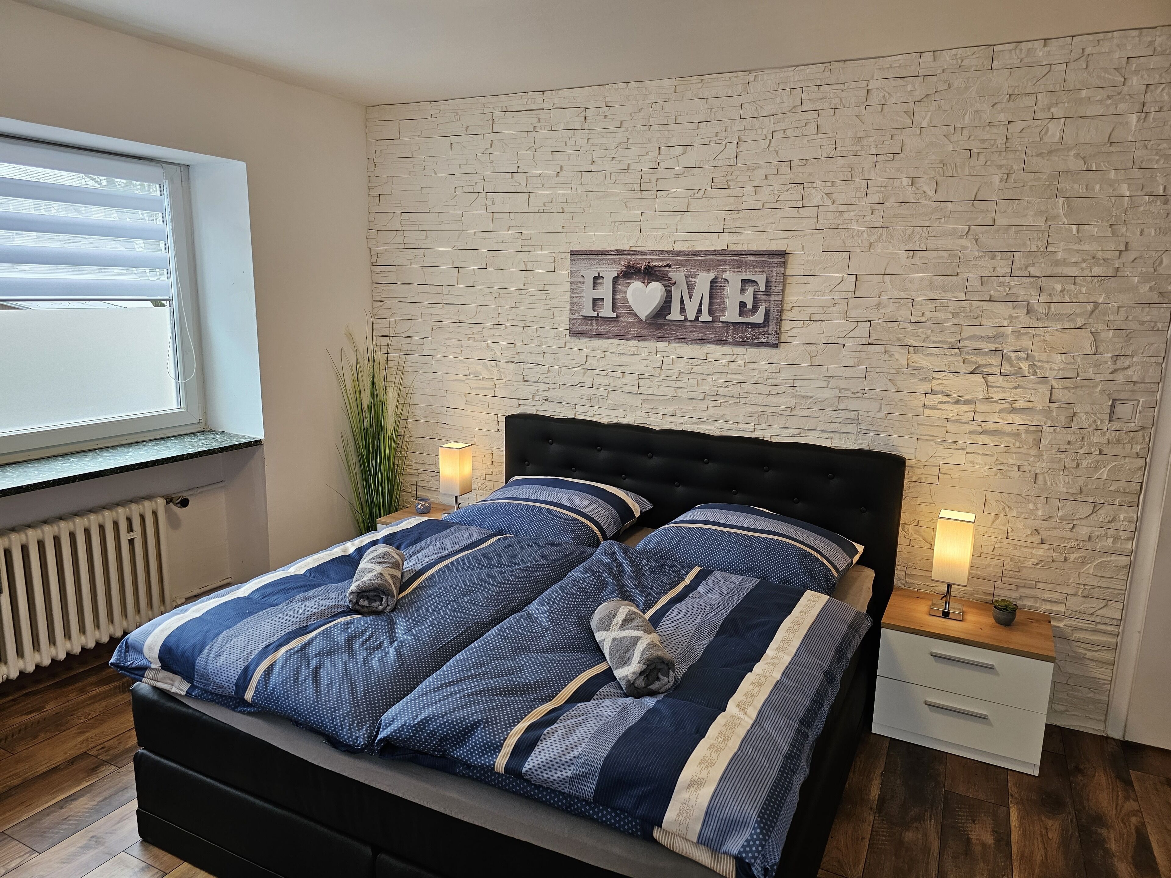 A bedroom with a stone wall, a bed with blue bedding, two bedside tables with lamps, and a window with blinds.