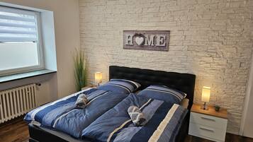 A bedroom with a stone wall, a bed with blue bedding, two bedside tables with lamps, and a window with blinds.