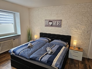 Een slaapkamer met een stenen muur, een bed met blauw beddengoed, twee nachtkastjes met lampen en een raam met zonwering.