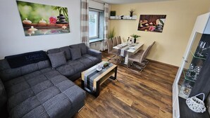 A modern living room with a grey sofa, a dining table with chairs, wooden flooring, and wall art depicting nature scenes. - Bright and modern apartment on the ground floor in the center (Kassel)
