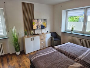 Ein Schlafzimmer mit Bett, Kommode, Sessel, Fernseher und Fenster mit Blick ins Grüne.