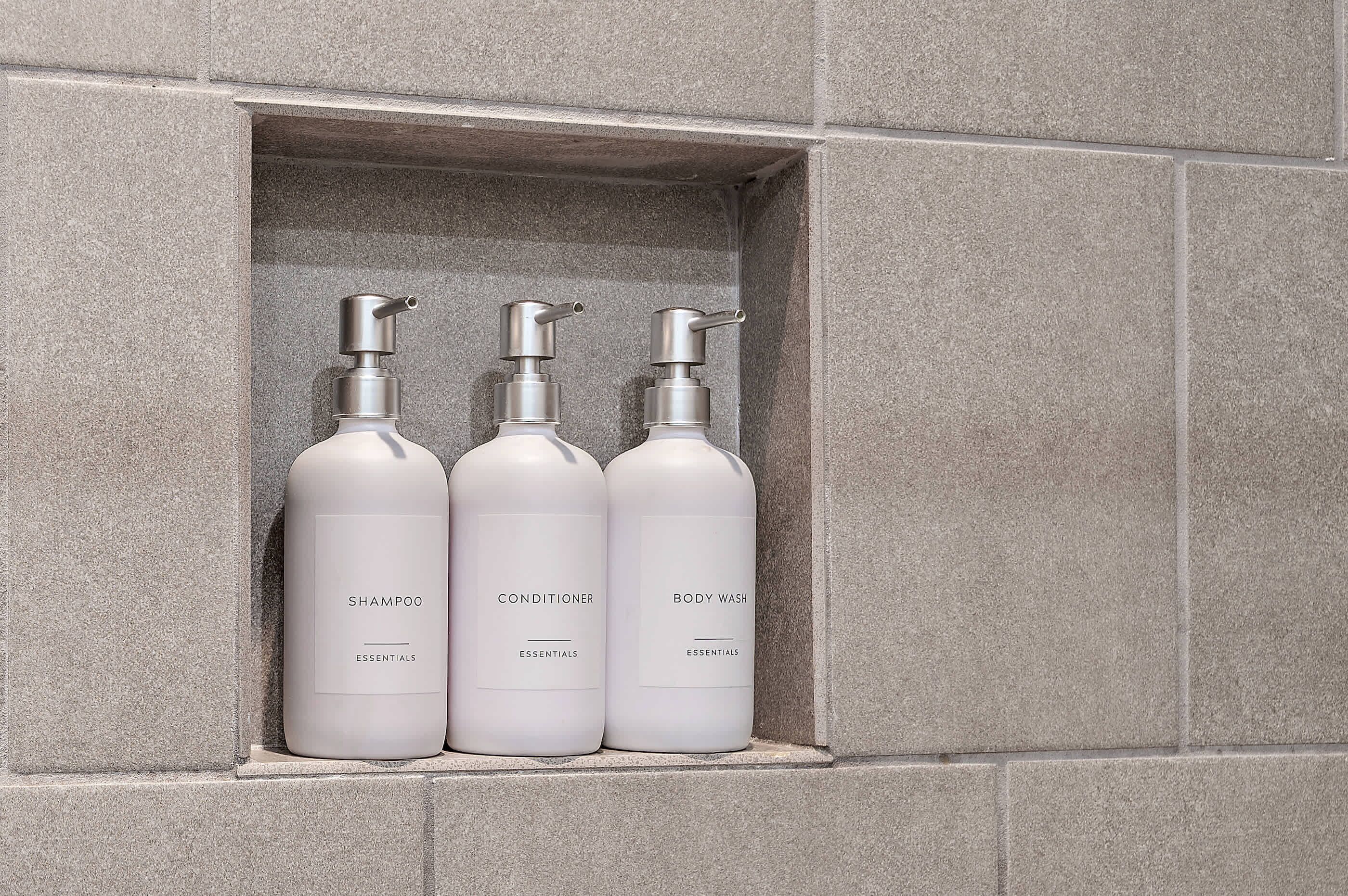 Three bottles of shampoo, conditioner, and body wash in a recessed shelf against a textured wall.