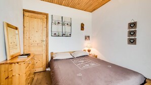 A bedroom with a wooden ceiling, a bed with a brown bedspread, a wooden dresser, a door, and wall-mounted decorations including a framed picture and heart-shaped frames.