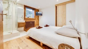A modern bedroom with a wooden floor, a large bed with white bedding, a glass shower enclosure, a wooden headboard, and a mounted TV.