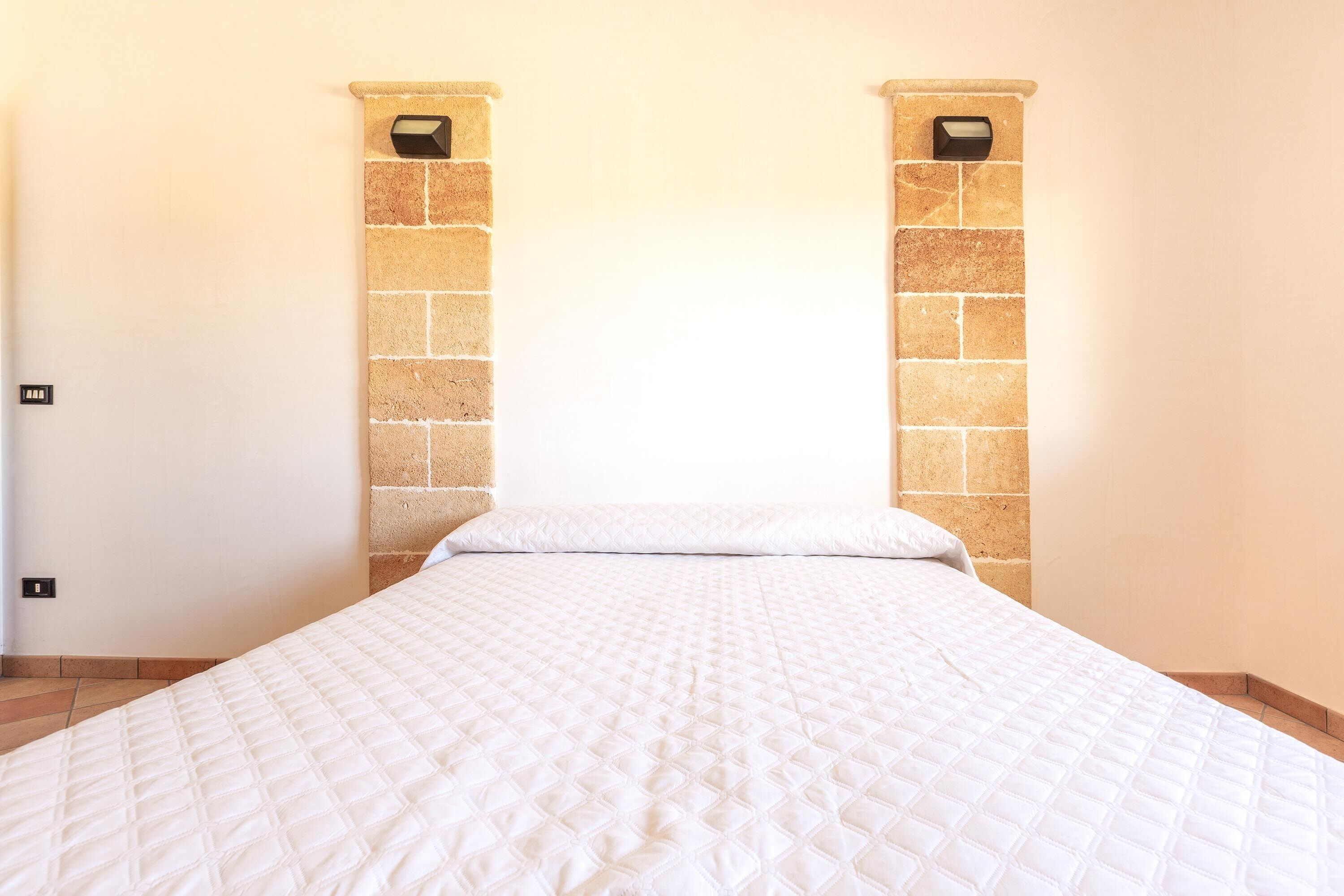 Un lit bien fait dans une pièce dont un mur est en pierre et où se trouve un interrupteur sur le mur.