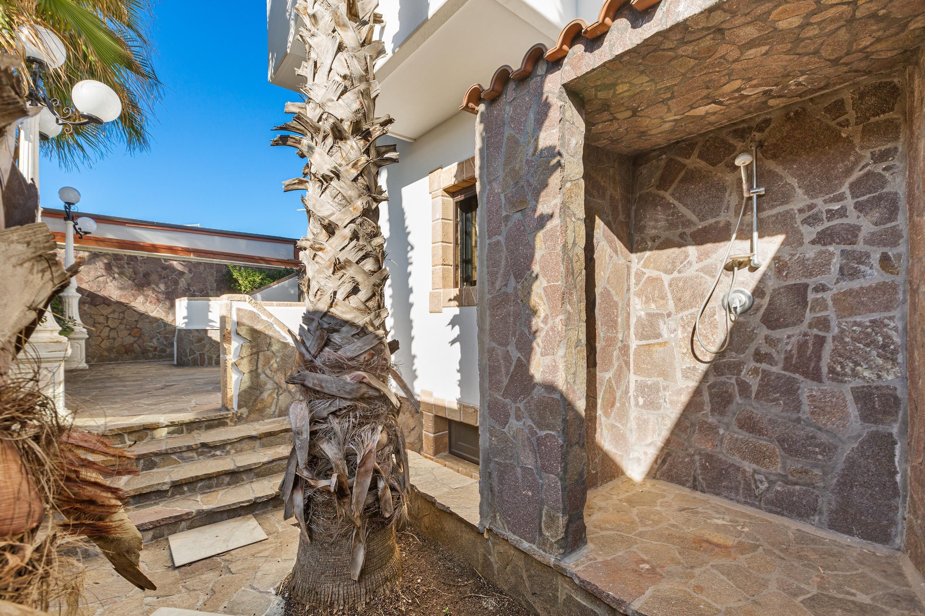 A stone-walled outdoor shower area adjacent to a white building with a stone staircase leading to a covered patio.