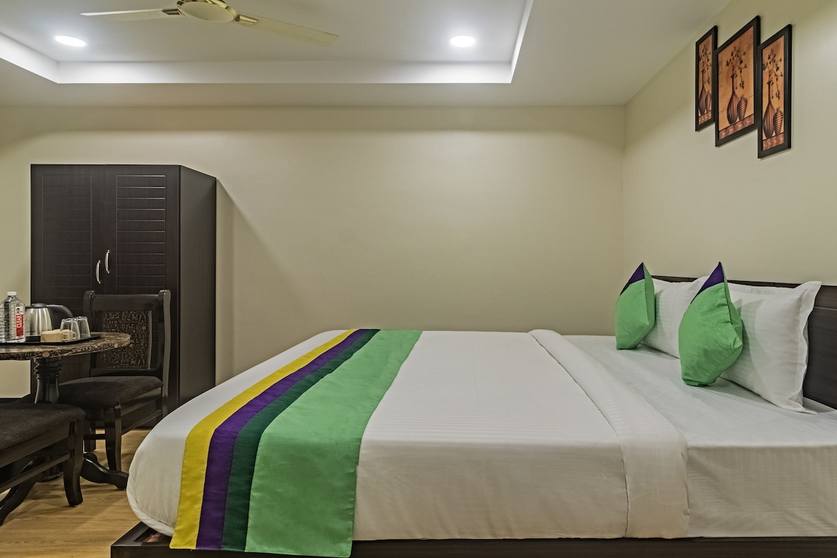 A neatly made bed with a white and green striped bedspread, a dark wooden headboard, and two green pillows. There is a small table with a tray of items, a dark wooden wardrobe, and a ceiling fan.
