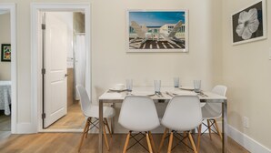 Een eetgedeelte met een tafel voor vier personen, witte stoelen en een schilderij met lijst erboven.
