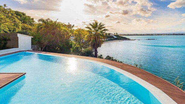 Une piscine dotée d’une terrasse en bois, entourée d’une végétation luxuriante et offrant une vue sur une mer calme.