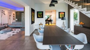 Un espace de salle à manger moderne avec une longue table, des chaises blanches et un piano en arrière-plan.
