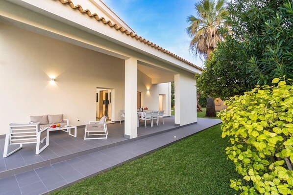 Un patio techado al aire libre con zona de comedor, mobiliario blanco y césped bien cuidado.