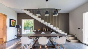 Une cuisine moderne avec un plan de travail foncé, des chaises blanches et un escalier agrémenté de luminaires suspendus noirs.