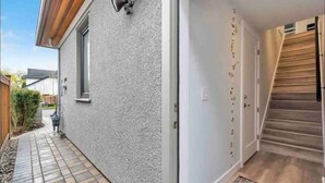 L’extérieur d’une maison moderne, avec une terrasse carrelée, un mur extérieur blanc et un escalier en bois menant à une porte.