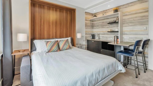 Une chambre moderne avec un grand lit, une tête de lit en bois, une petite kitchenette et un bureau avec deux chaises.