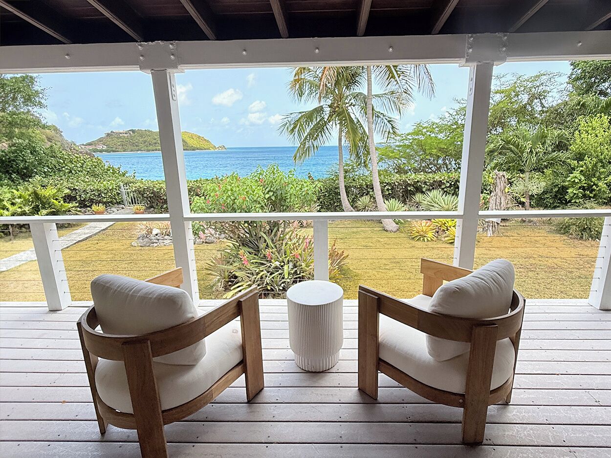 A wooden deck with two cushioned chairs and a small round table, overlooking a tropical landscape with a clear view of the ocean and hills.