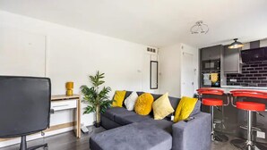 Una sala de estar moderna con un sofá seccional, un escritorio y un televisor. Se observan taburetes rojos y una cocina al fondo.