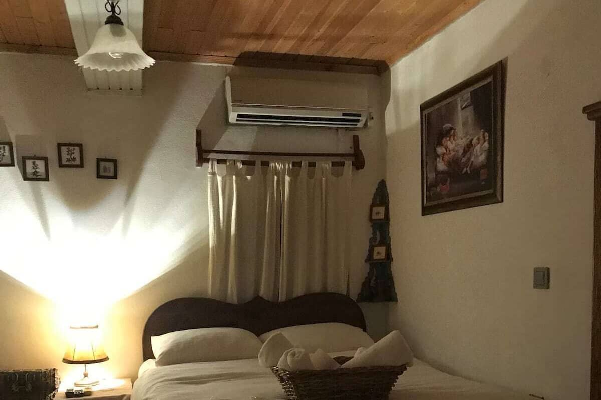 A bedroom with a bed, bedside tables, an air conditioning unit, a lamp, curtains, and a framed picture on the wall.