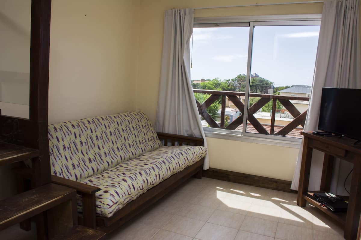 A room with a sofa, a TV on a wooden stand, and a window with a view of buildings and trees.