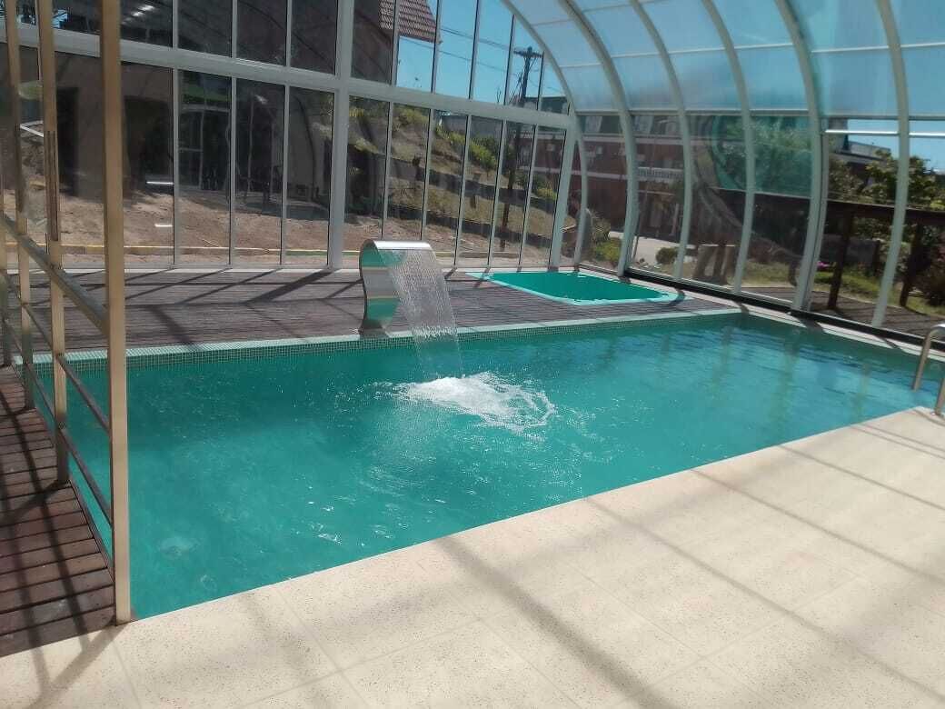 An indoor swimming pool with a clear glass enclosure, a diving board, and a small waterfall feature.