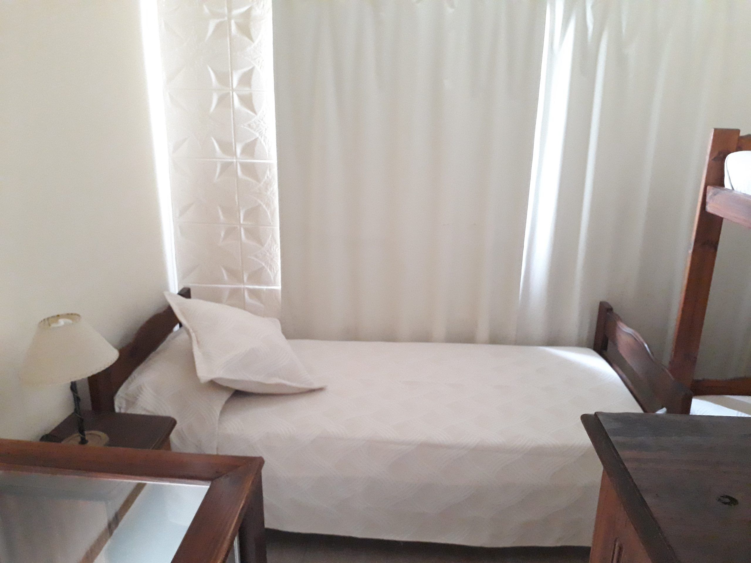 A small, neatly made bed with a white bedspread and a single pillow. There is a wooden nightstand with a lamp on it to the left of the bed. A wooden dresser is positioned to the right of the bed.