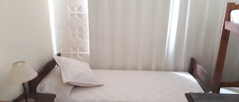 A small, neatly made bed with a white bedspread and a single pillow. There is a wooden nightstand with a lamp on it to the left of the bed. A wooden dresser is positioned to the right of the bed.
