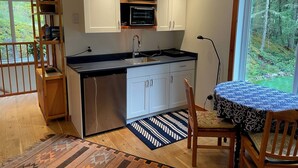 A compact kitchen with white cabinets, a built-in microwave, and a dishwasher. There's a dining area with a table and chairs, a patterned rug, and a view of a forest through a large window. - Modern studio. (Salt Spring Island)