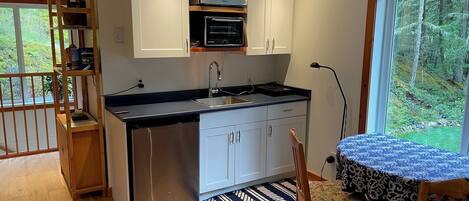 A compact kitchen with white cabinets, a built-in microwave, and a dishwasher. There's a dining area with a table and chairs, a patterned rug, and a view of a forest through a large window.