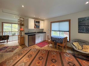 A compact kitchen with white cabinets, a microwave, and a sink. There is a dining area with a table and chairs, and a large window with blinds.