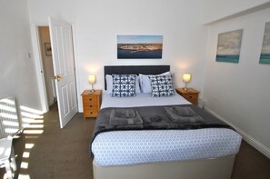 A neatly made bed with patterned bedding, two bedside tables with lamps, a door leading to another room, and a wall-mounted painting. - Sea Whisper- lovely home in charming village (Pittenweem)