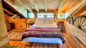 A cozy attic bedroom with a wooden floor, a bed with a patterned blanket, a small table with a lamp, and a window with a view.