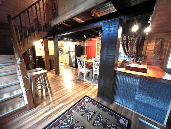 A rustic interior with a wooden staircase, a dining area with white chairs, and a kitchen with wooden cabinets.