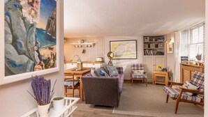 A cozy living room with a sofa, chairs, a coffee table, and a dining area with a table and chairs. There is a bookshelf with books and decorative items, and a large painting on the wall. - East Pier View- stunning shorefront home (Saint Monans)