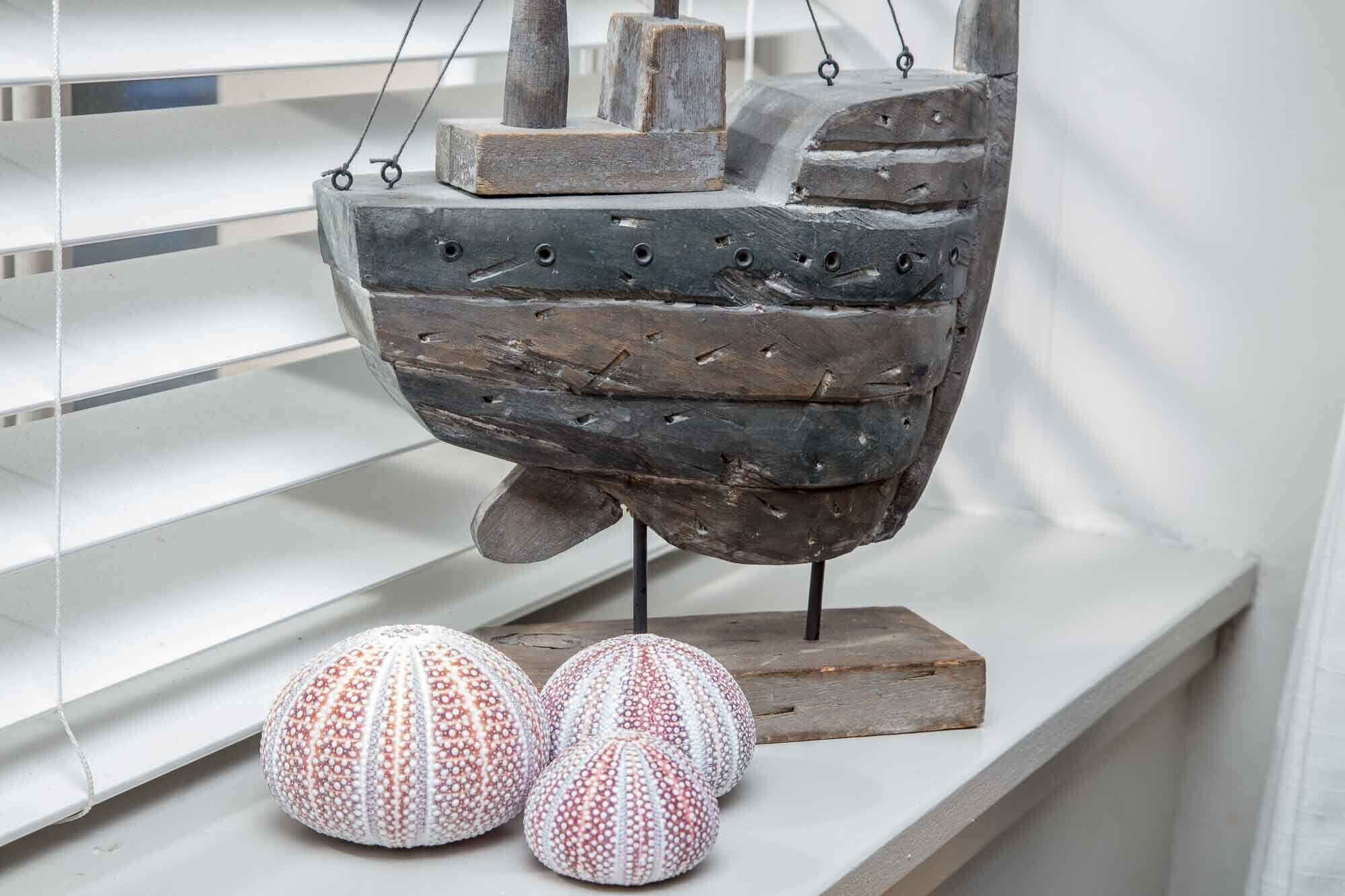 Een rustieke schelpkunstwerk met een houten voetstuk, twee bolvormige objecten ernaast, en een witte plank in de achtergrond.