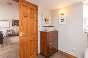 Una habitación con puerta de madera, un aparador con cajones, una mesita con una cesta y dos cuadros en la pared.