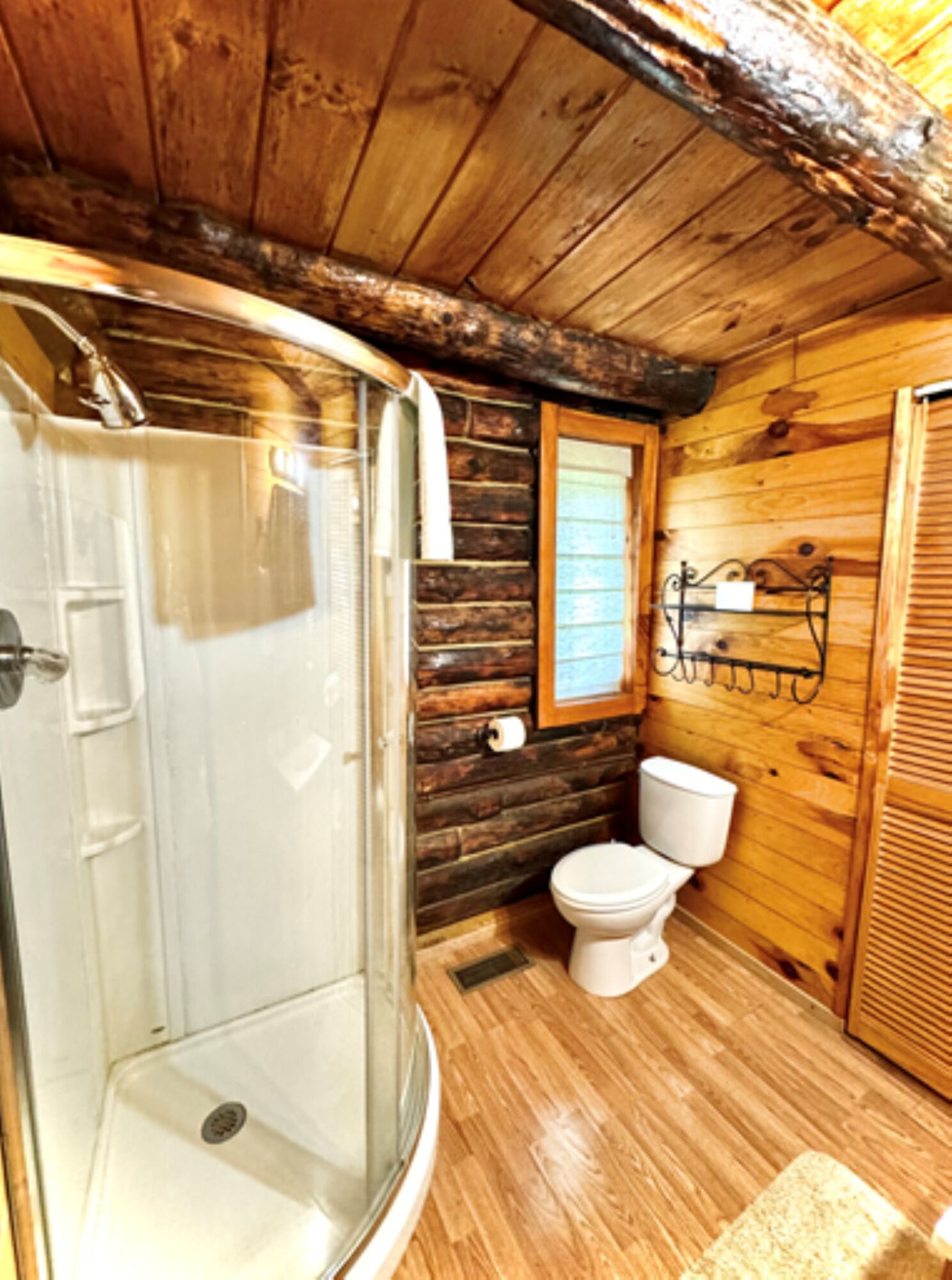 Een houten badkamer met een glazen douchecabine, toilet en raam.